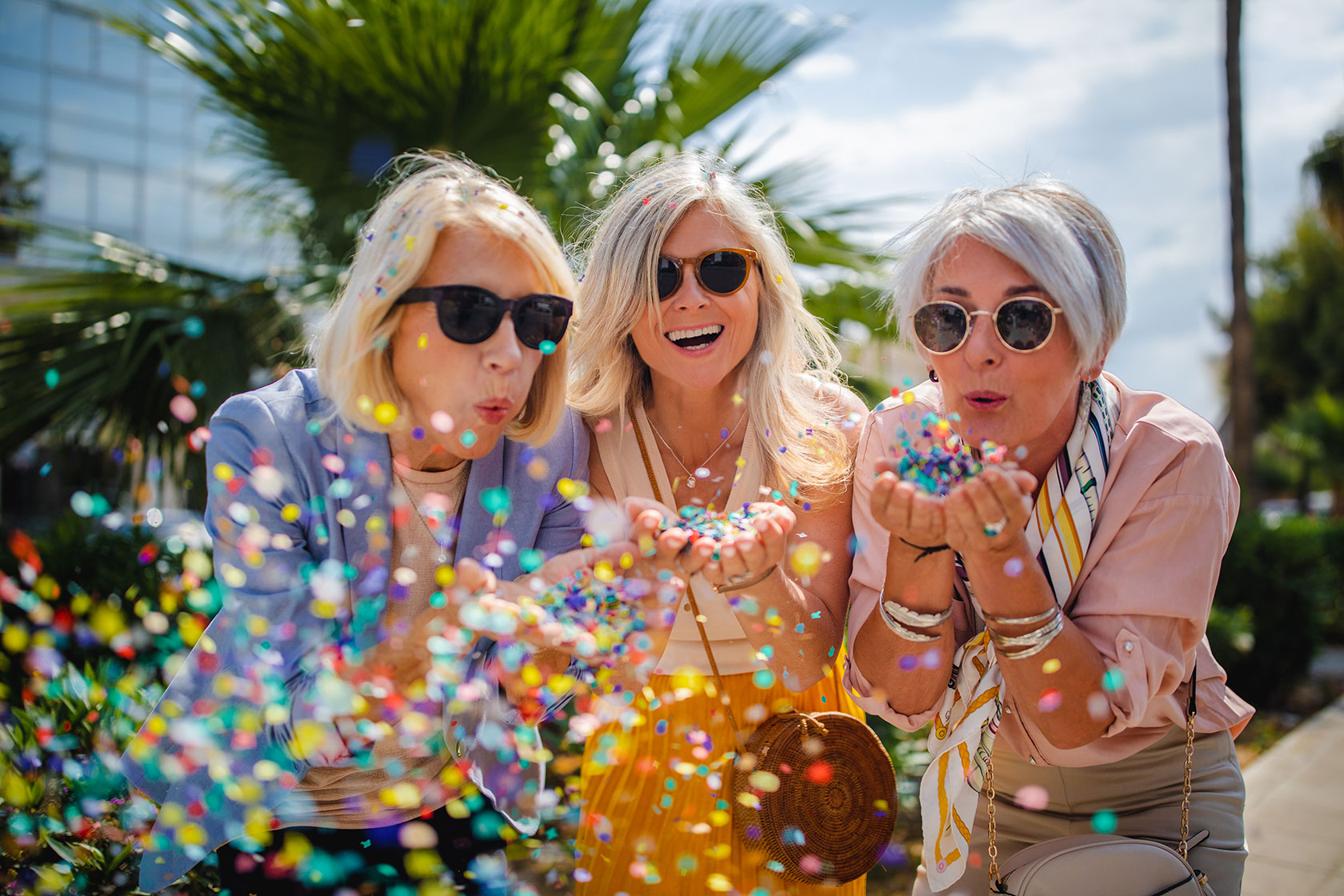 Three older women in sunglasses blowing confetti 